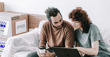 Couple sitting in a room with moving boxes and a wrapped sofa looking at a computer trying to plan a move